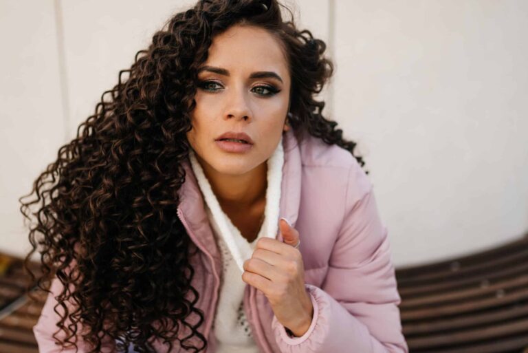 A brooding curly woman in a white sweater sits outside, close-up, looking to the side.