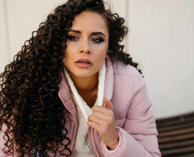 A brooding curly woman in a white sweater sits outside, close-up, looking to the side.