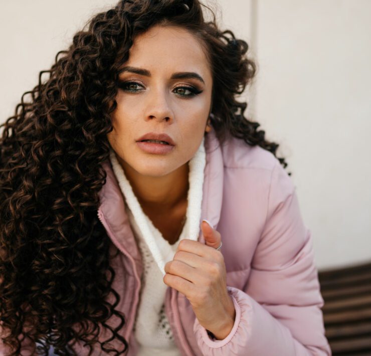 A brooding curly woman in a white sweater sits outside, close-up, looking to the side.