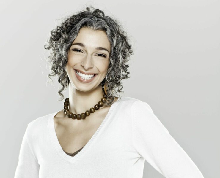 A woman with curly gray hair stands with her hands on her hips while smiling