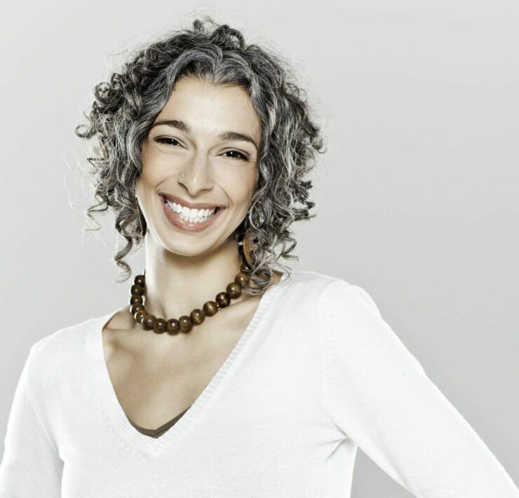 A woman with curly gray hair stands with her hands on her hips while smiling